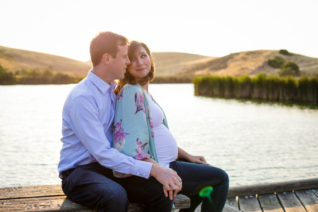 husband and expectant wife sitting next to water at sunset
