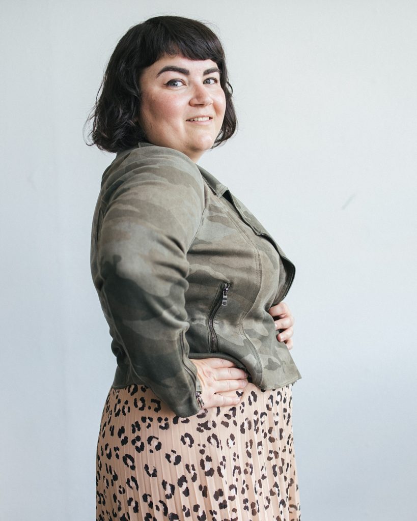 A confident family photographer with dark hair poses for a portrait while mixing a camouflage print jacket and leopard print skirt