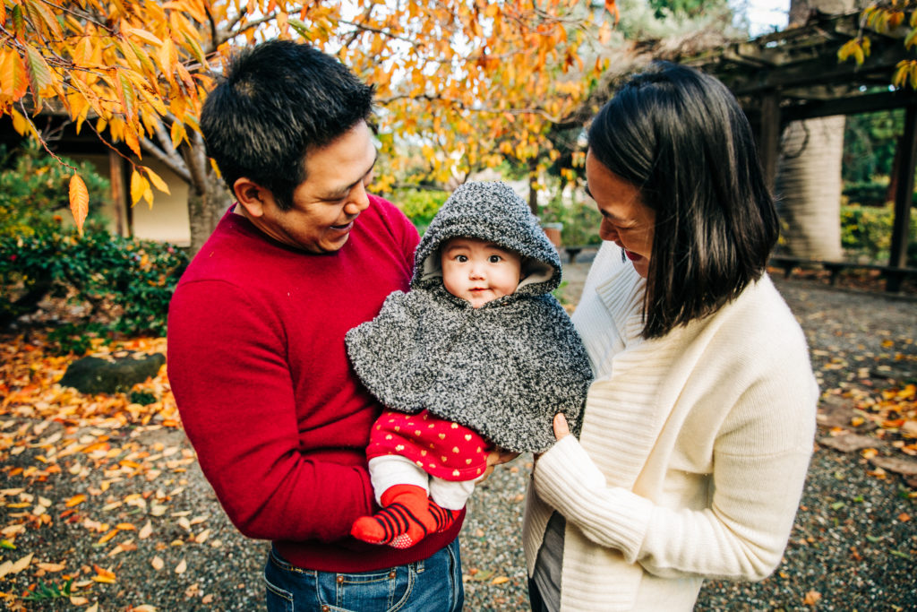A man in a red sweater and woman in a white cardigan hold their cute child during an outdoor fall family photography session