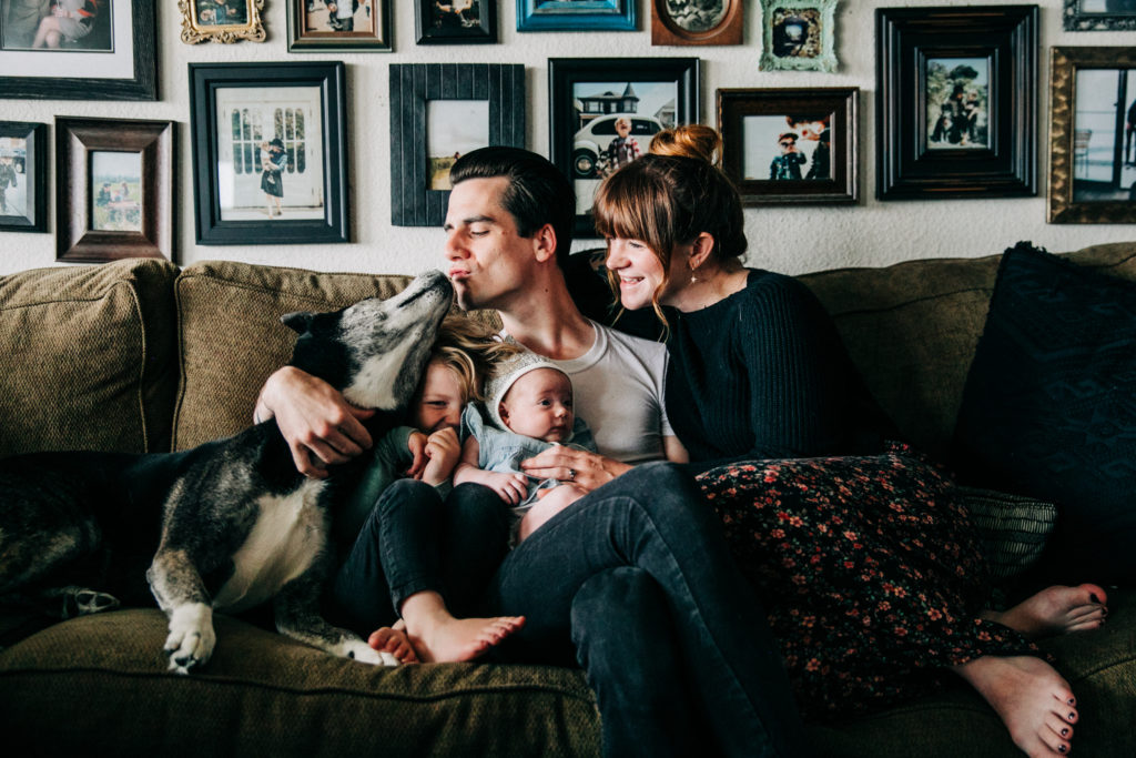 A trendy couple sits on the couch in front of a collage wall with their baby, son and dog for a family photo session