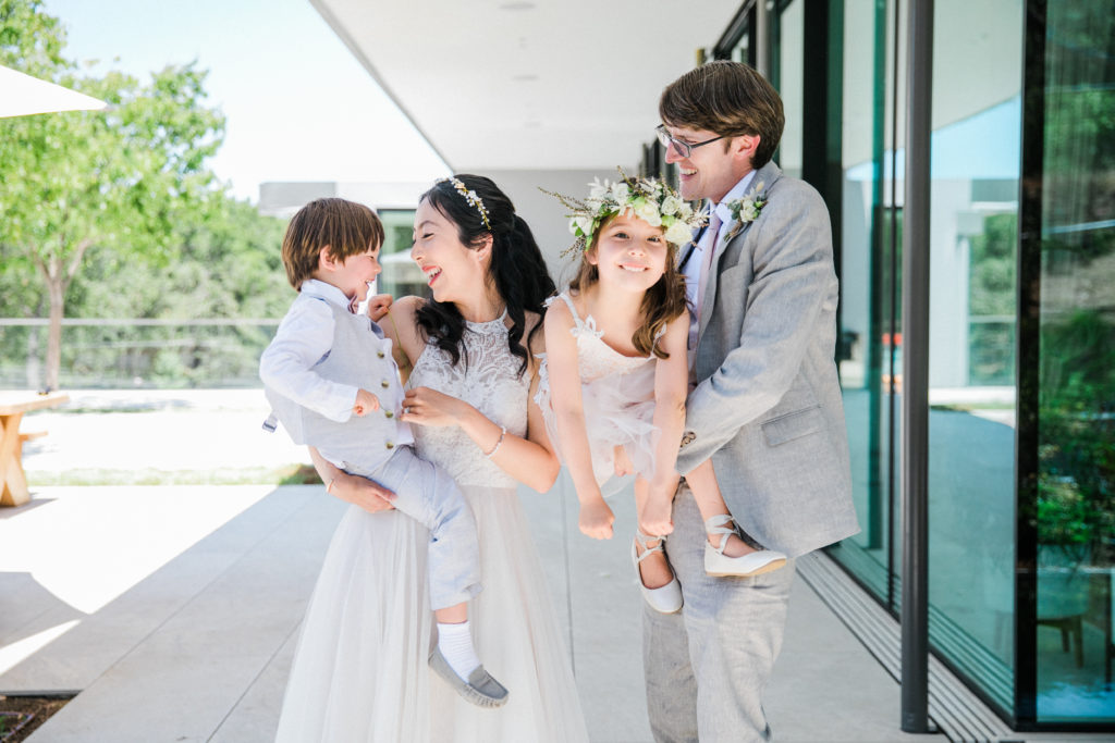 husband and wife in suit and wedding dress holding kids in wedding attire