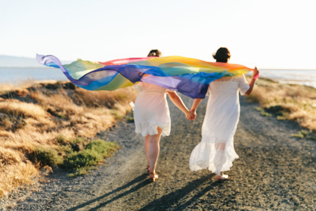 two women in white dresses walking with rainbow flag