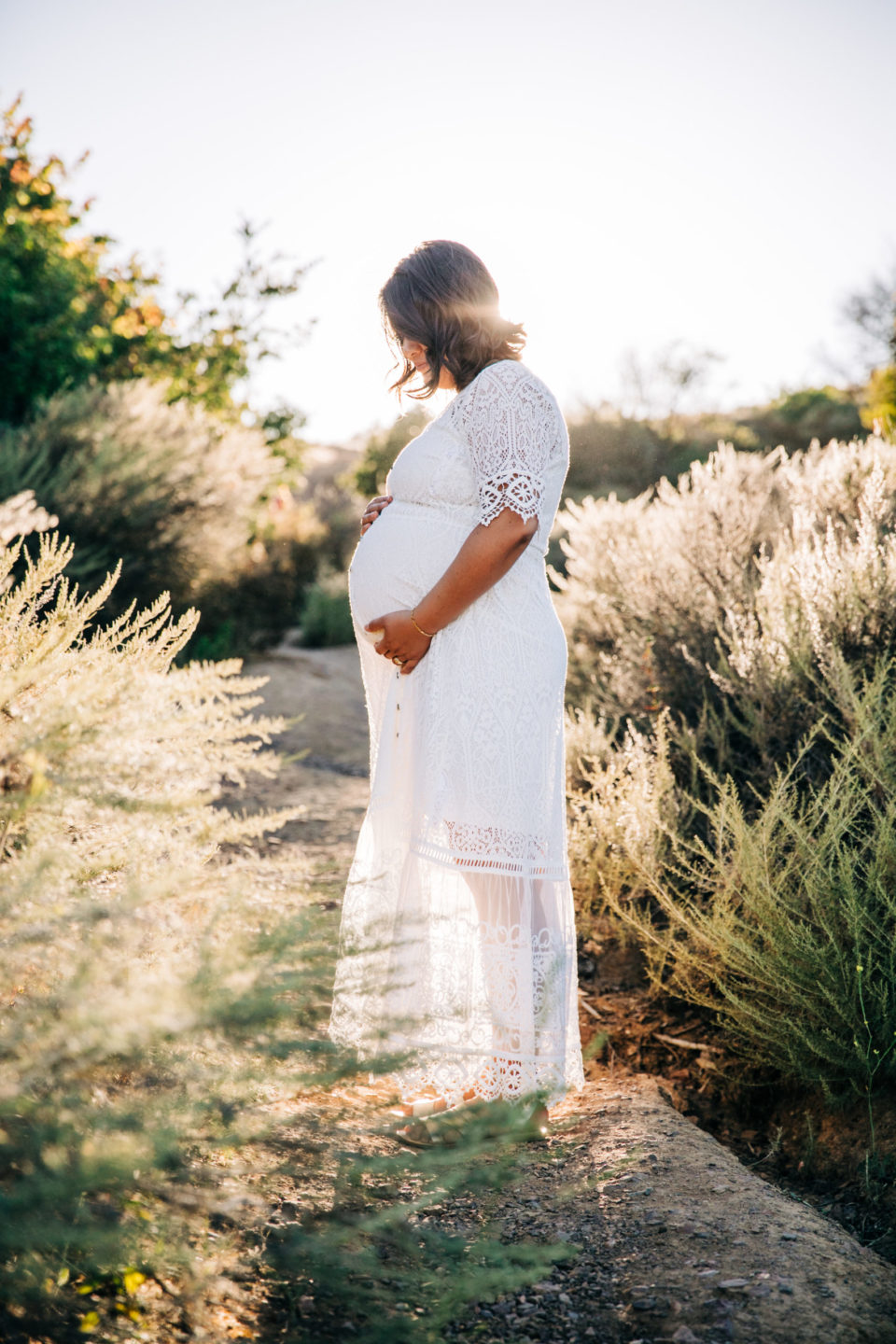 pregnant woman in long white dress backlit in nature