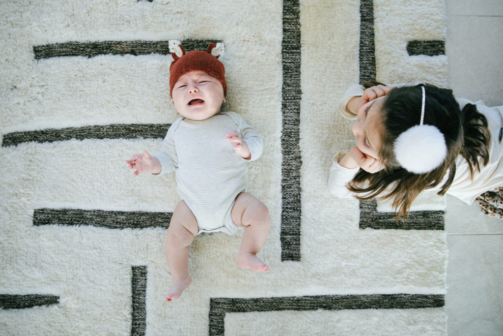 birds eye view of baby crying on rug while older sister watches from the side