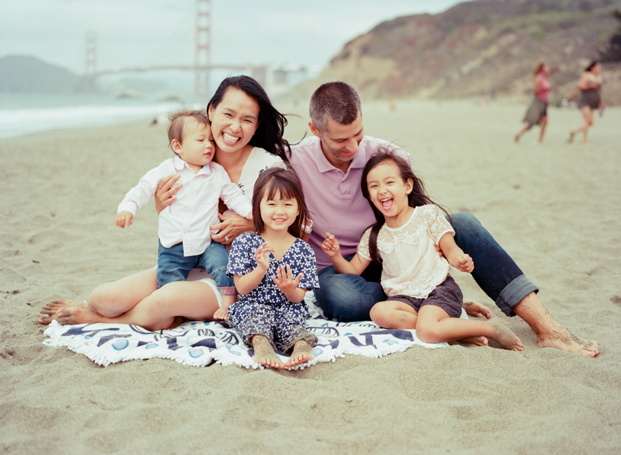 family of five sitting on a blanket at baker beach with the golden gate bridge in the background