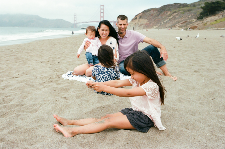family sitting on beach with one daughter pouring sand on her legs