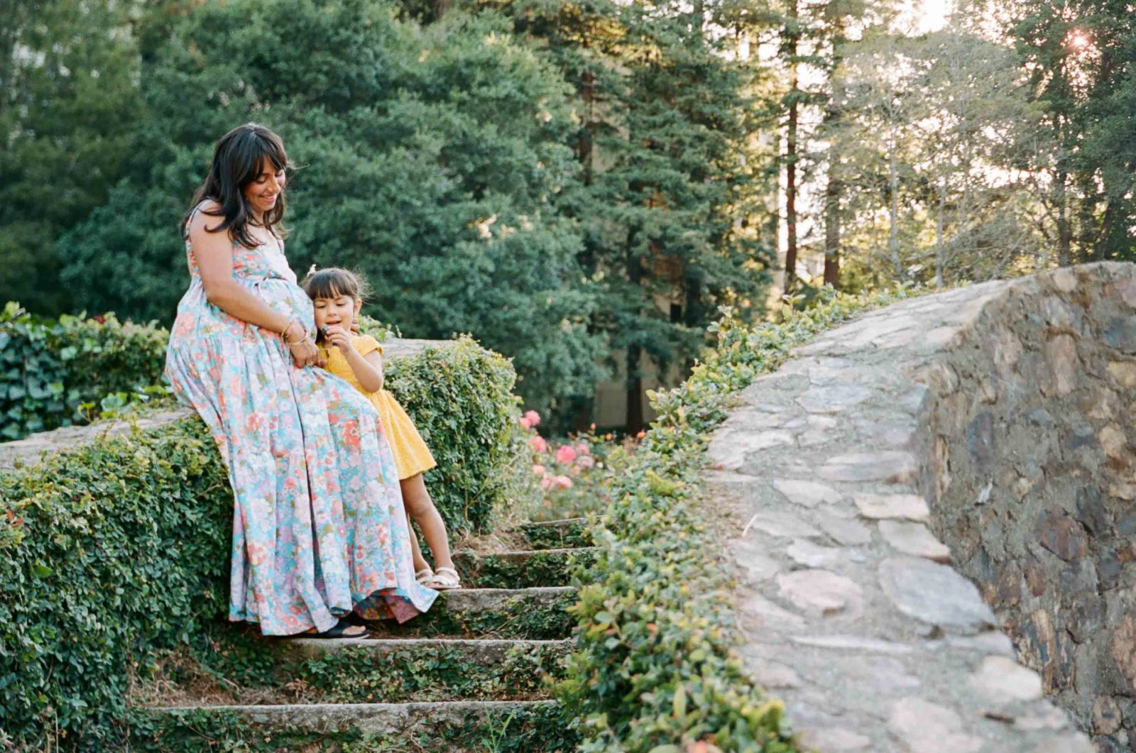 pregnant mama and toddler daughter sitting on stone wall covered in ivy