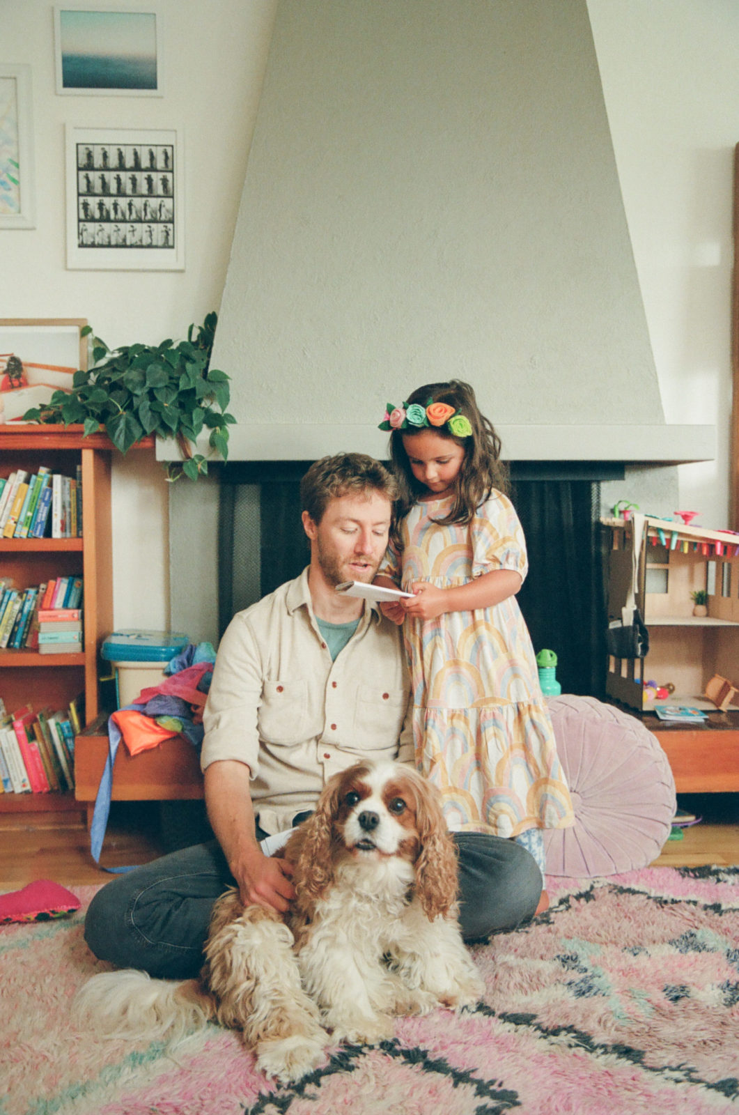 photo of a dad sitting cross legged on the living room floor with the family dog while young daughter shows him something in a book