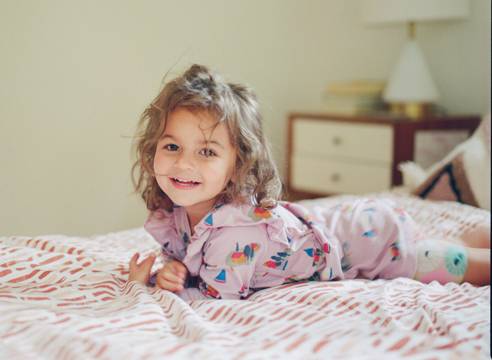 photo of a toddler girl with messy hair lying tummy down on the bed smiling at the camera