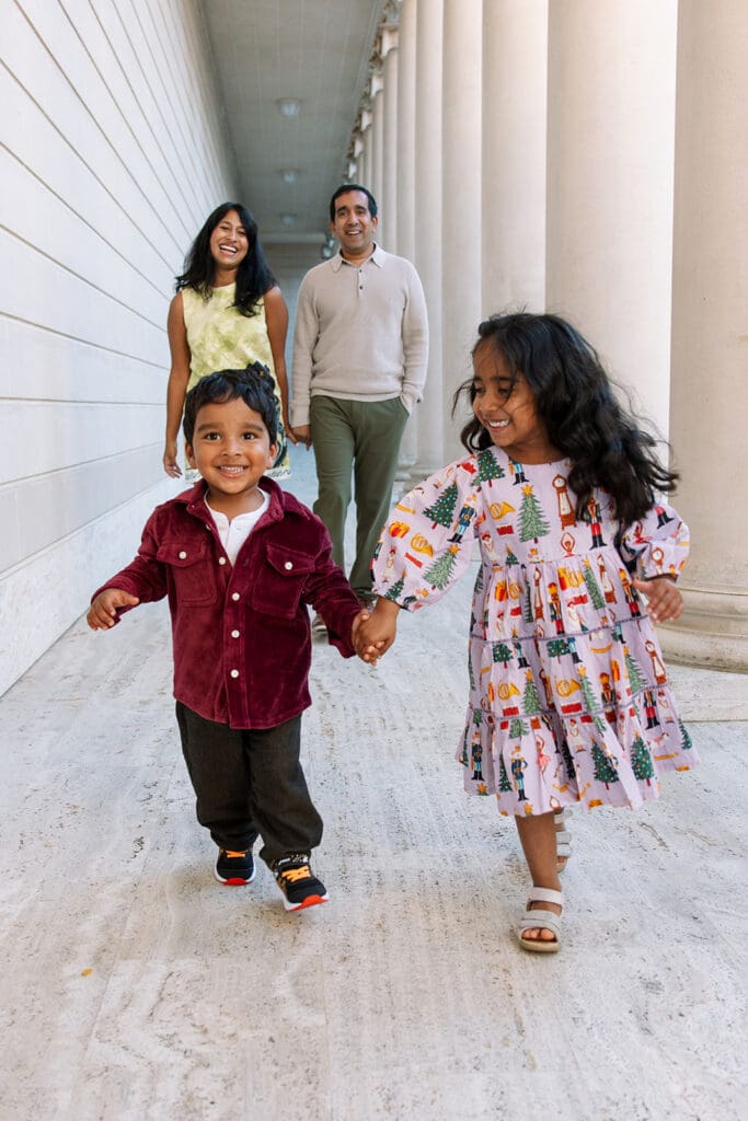 young brother and sister holding hands and running towards the camera while mom and dad smile and walk toward them from behind
