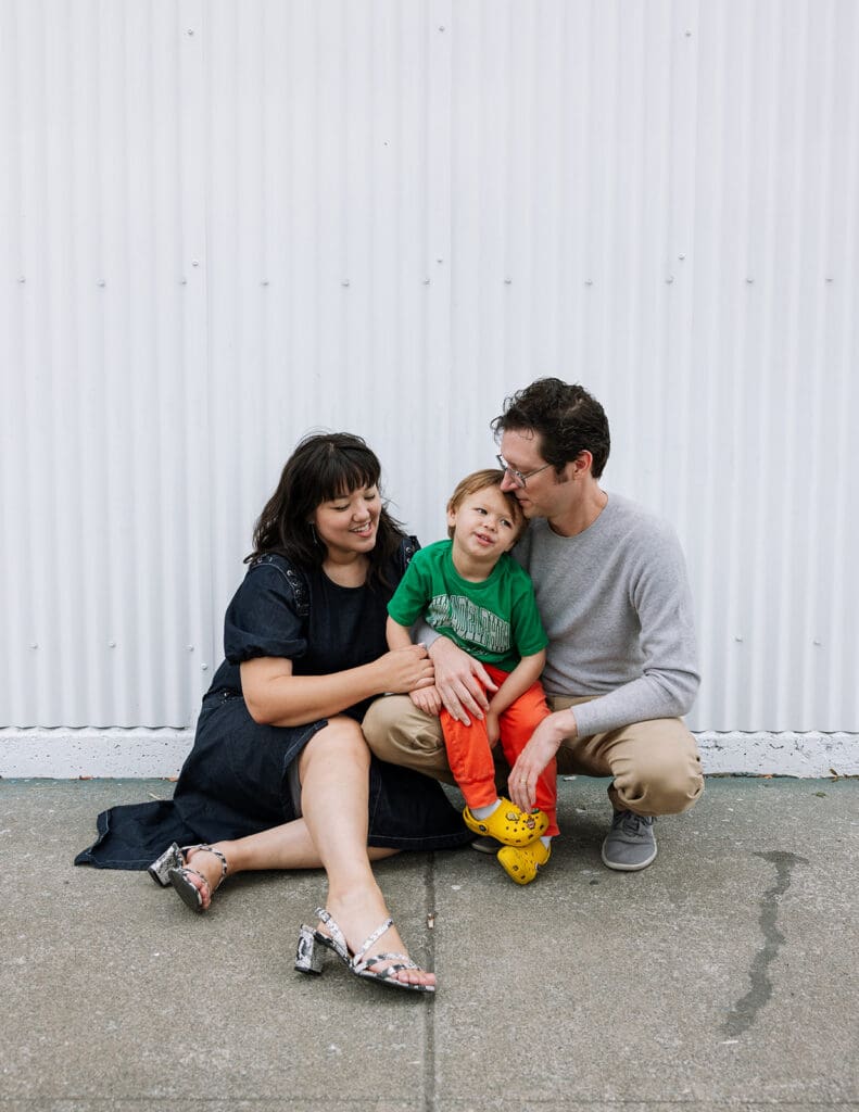 family of three sitting on sidewalk in front of white corrugated wall