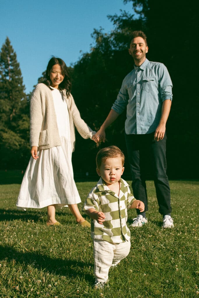 mom in white dress and beige cardigan holding hands with dad in chambray shirt and jeans standing behind toddler son 
