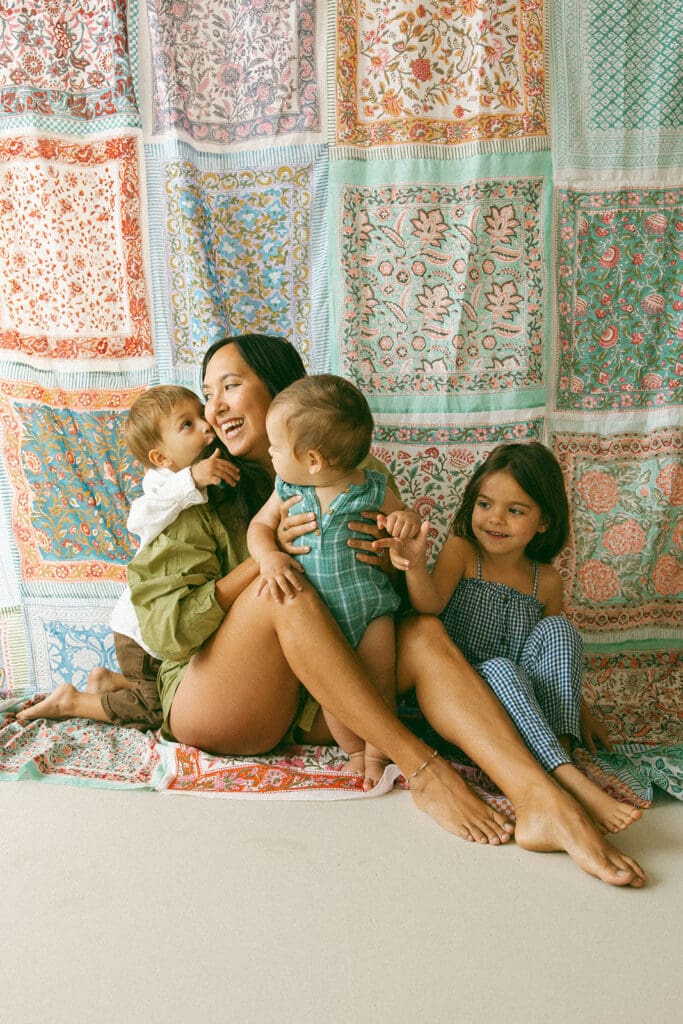 Mom in green coordinating set holding baby boy while toddler boy hugs her from behind and young daughter sits next to her smiling