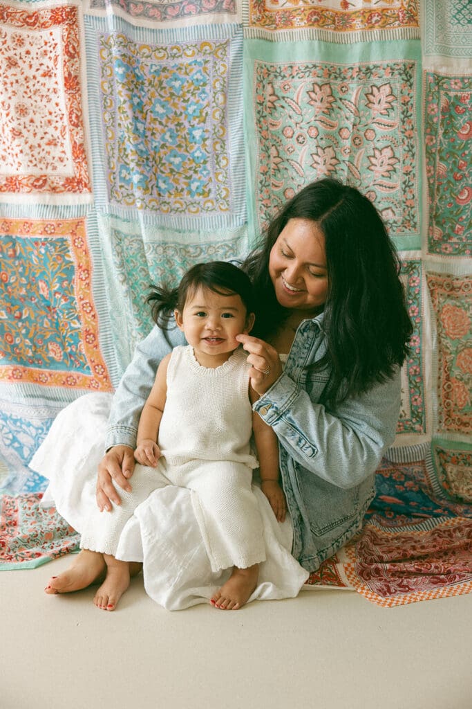 mom in denim jacket and white dress sitting cross legged with toddler girl in white romper on her lap in front of a handmade scarf backdrop