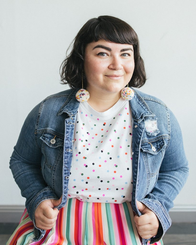 A woman with dark hair smiles at the camera while wearing fun polka dot earrings, a colorful polka dot shirt, a striped skirt and a denim jacket, showing tips for mixing patterns