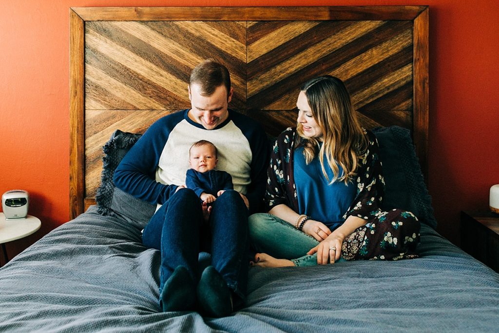 A man and woman sitting on their bed with their baby, posing for lifestyle newborn pictures with Bay Area newborn photographer Hi + Hello Photo