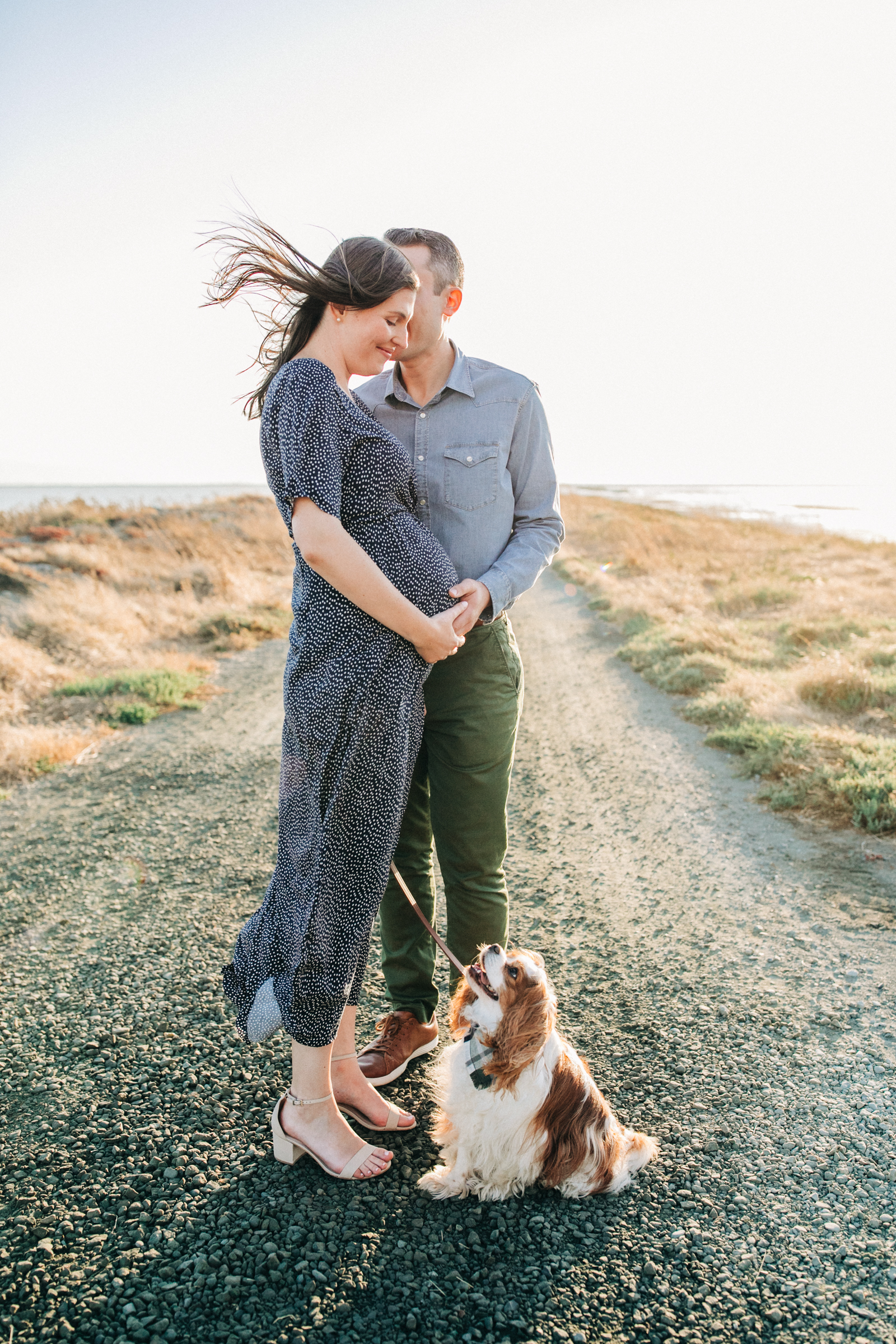 A pregnant couple stands together with their dog on a dirt road by the San Francisco waterfront
