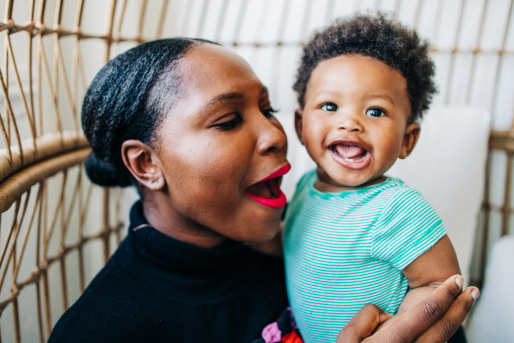 A beautiful mother wearing red lipstick holds her son while he smiles at the camera during an at-home family photography session in San Francisco 