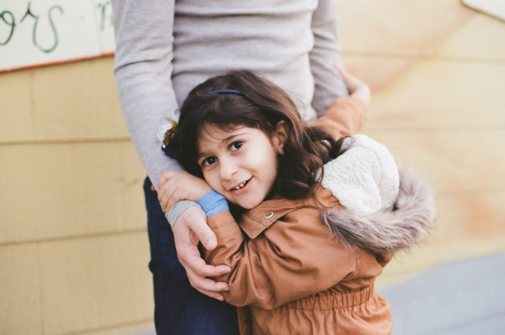 a young girl with dark hair and a brown coat smiles at the camera while hugging her mom while having outdoor family photos taken