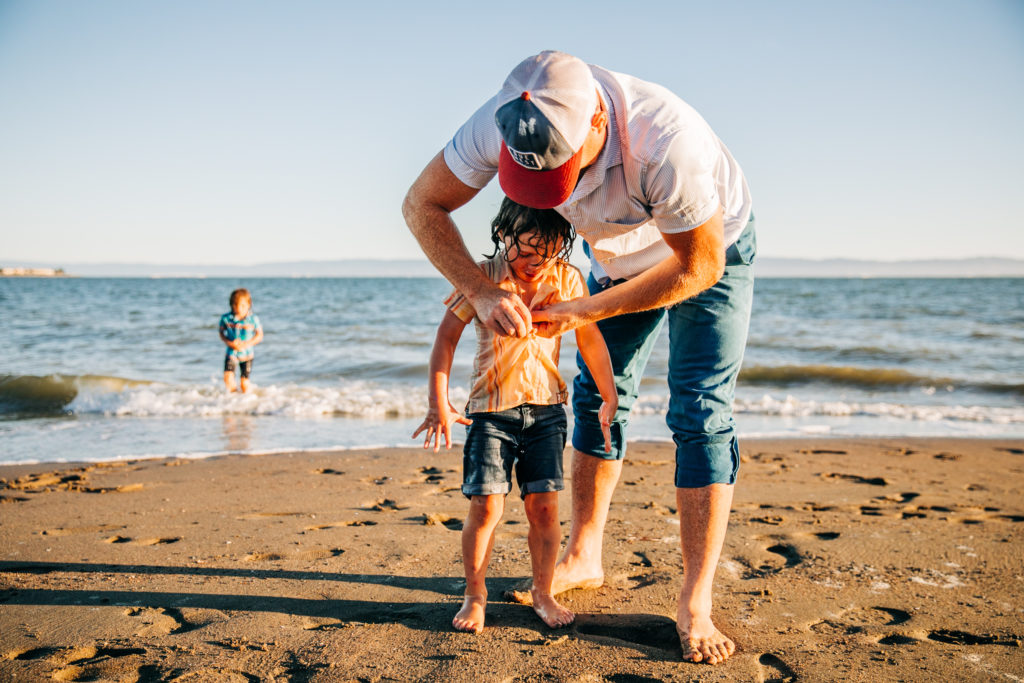 A father helps his son with his shirt buttons at the beach after jumping in the water for family photos in the Bay Area
