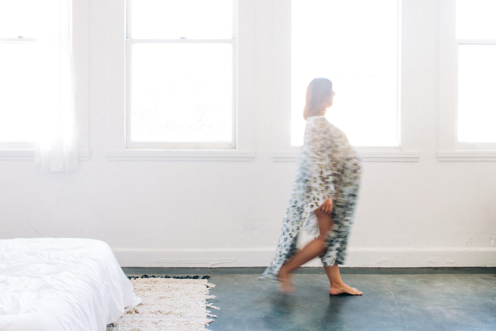 A pregnant woman wearing a flowing dress walks in front of a window during an indoor maternity session with Hi + Hello Photography