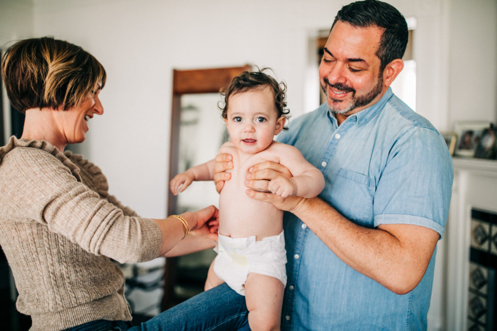 A father holds his baby while his wife puts on a diaper during a lifestyle family photography session at home