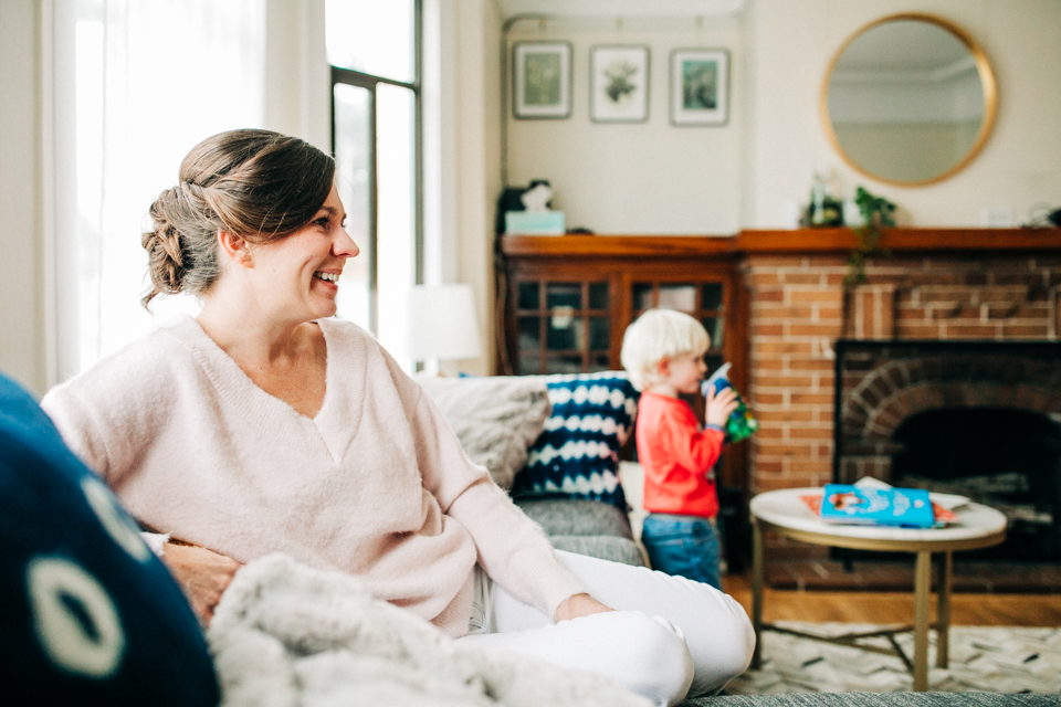 A woman in a pink sweater sits on her couch and smiles while her toddler plays in the background during an indoor family session in the Bay Area