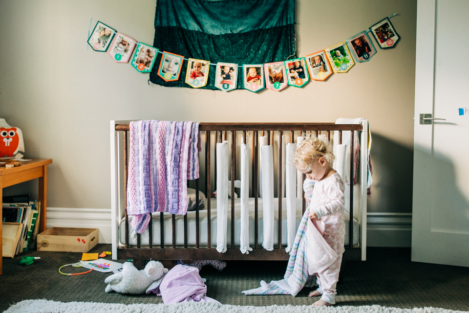 A toddler walks in front of her crib while carrying her blanket during a lifestyle family photo session with Bay Area family photographer Hi + Hello Photography