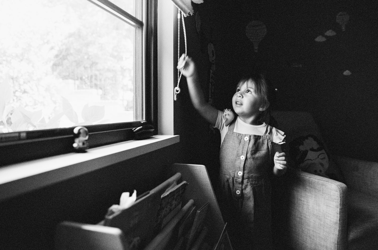 black and white Photo of a young girl pulling on the cord to open the blinds of her bedroom window