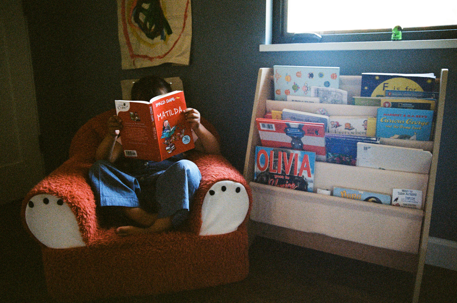 color film photo of young girl sitting in a bear chair reading a paperback copy of the book Matilda